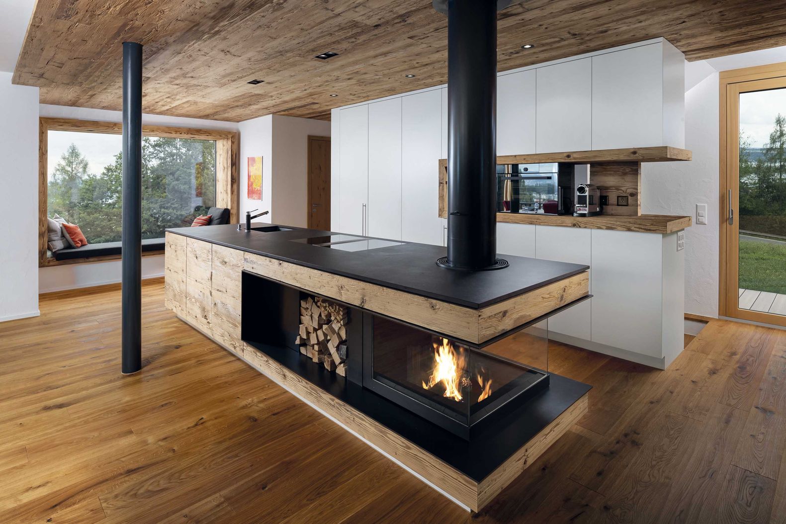 Die mit Massivholz verkleidete Feuerstelle ist im Wohnbereich platziert und doch so konzipiert, dass im Arbeitsbereich der Küche möglichst wenig Hitze entsteht. Wirth & Schmid AG.