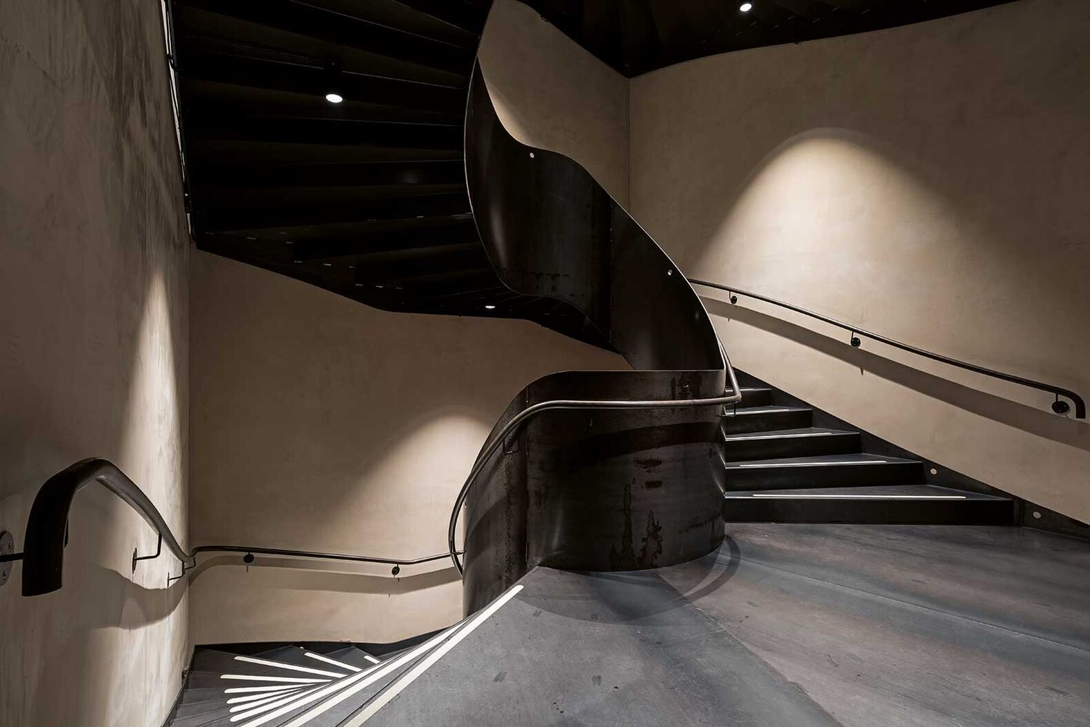 Die grosszügige Wendeltreppe aus Schwarzstahl füllt das Treppenhaus vollständig aus. Ihre Dimensionen sind aussergewöhnlich: Mit bis zu drei Metern Breite beim An- und Austritt sowie einer Mindestbreite von eineinhalb Metern wirkt sie weniger wie eine blosse Treppe, sondern vielmehr wie ein skulpturales Element. Insgesamt wurden vier dieser Treppen übereinander realisiert, jede mit einem Gewicht von rund 13 Tonnen. Das dunkle Material wirkt durch die organisch geschwungene Form fliessend. Weswegen die Treppe trotz ihrer Masse nicht schwer oder sperrig wirkt, sondern geschmeidig den Raum einnimmt. Sie befindet sich im Hortus-Gebäude und gibt mehreren Personen zugleich Platz, um in das gewünschte Stockwerk zu gelangen. Ferdinand Hasler AG, www.hasler-treppen.ch
