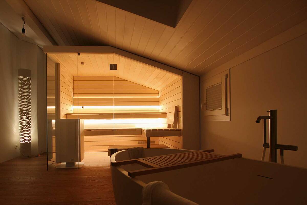 Die Sauna und die Sitzbänke wurden bei diesem Objekt an die Dachschräge angepasst. Eine gemütliche Atmosphäre schafft die indirekte, warmweisse LED-Beleuchtung. Die Glasfront lässt den Raum grosszügig wirken. Ging Saunabau AG.
