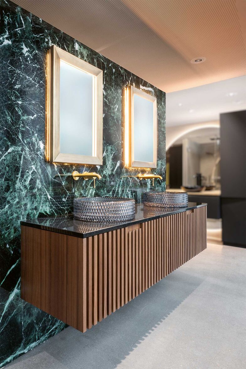 Dieser Bereich zeigt sehr schön, dass Möbel mit Lamellen ebenso mutig kombiniert werden können, wie minimalistischere Modelle. Das Badmöbel ist mit Nussbaum furniert und die vertikalen Leisten bestehen aus Massivholz. Anstelle eines Griffs gibt es einen Grifffalz an den Leisten. Mit den goldenen Akzenten durch die Armaturen und Spiegel harmoniert das dunkle Holz sehr schön und drückt Eleganz aus. Die Rückwand besteht aus poliertem Naturstein und die Abdeckung aus poliertem Granit. Der Glanz dieser Oberflächen spiegelt die Struktur des Glasaufsatzbeckens wieder und erzeugt eine fast opulente Atmosphäre, ohne zu schwer zu wirken. Schreinerei Fehlmann AG, www.schreinerei-fehlmann.ch